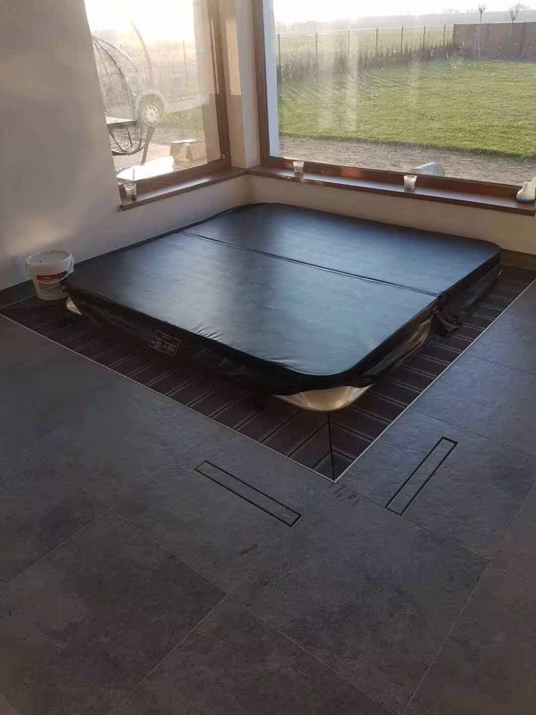 Domowe spa z dużym jacuzzi przykrytym czarną matą termiczną, usytuowane w rogu pokoju przy dużych oknach z widokiem na ogród.